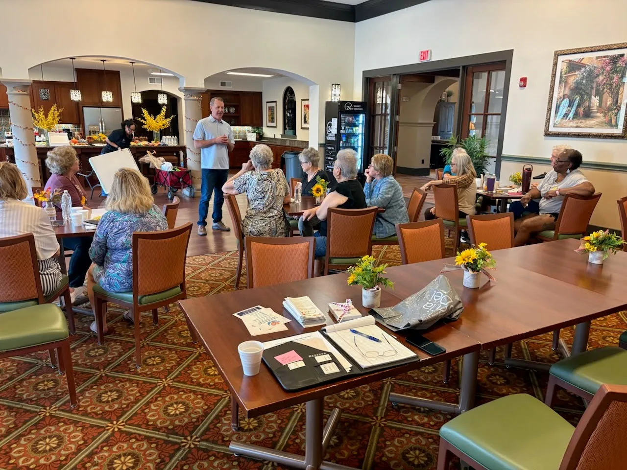 Stephen Nadalin standing and speaking to a group of residents seated at tables in a retirement home activity room in Webster